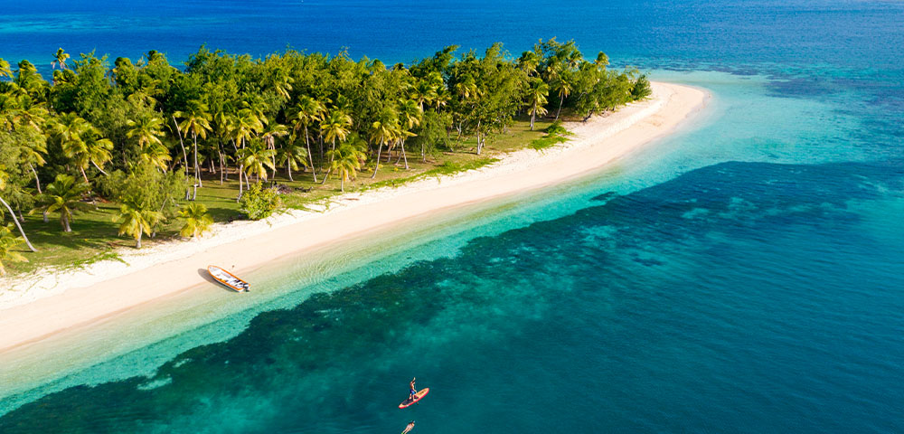 Aerial view of a palm-fringed beach in Fiji's Yasawa Islands with a paddleboarder on turquoise water
